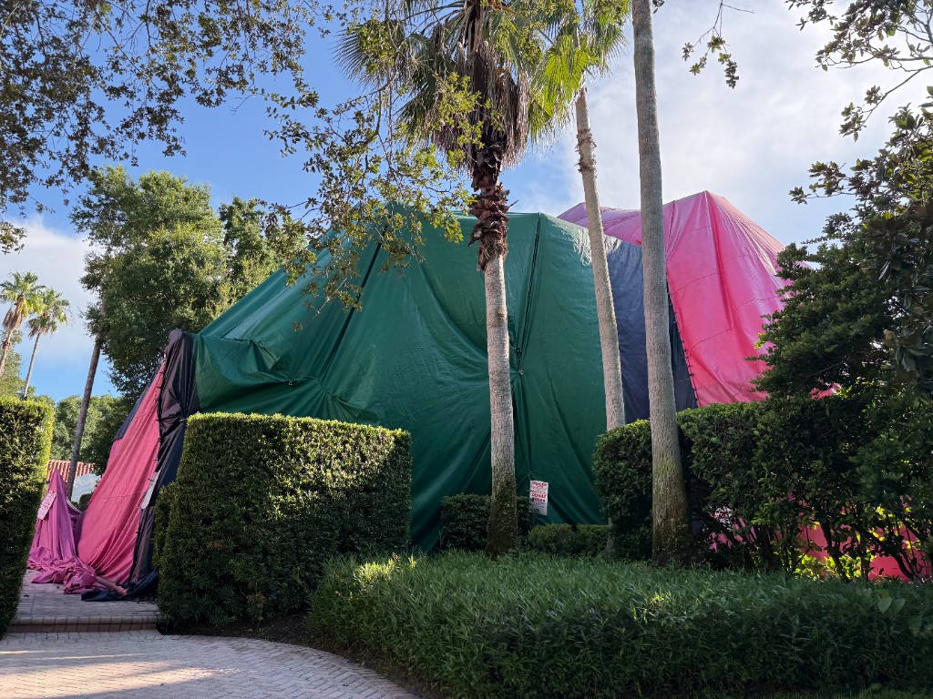 House tented with green and pink fumigation tarps, palms, and a posted warning sign